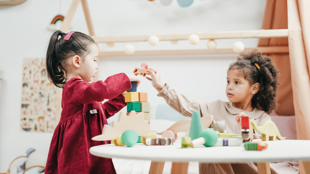 Two young girls enjoying playtime with wooden toys indoors in a warm, colorful playroom.