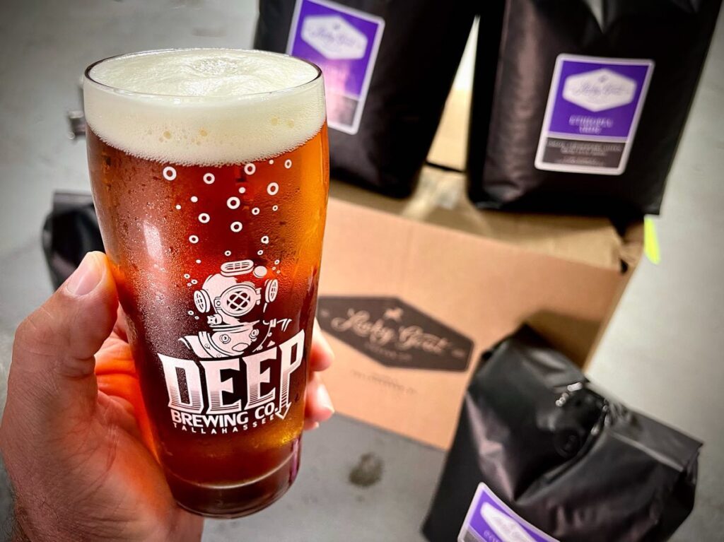 Man's hand holding a glass of beer with the Deep Brewing Co logo, with Lucky Goat coffee packages in the background